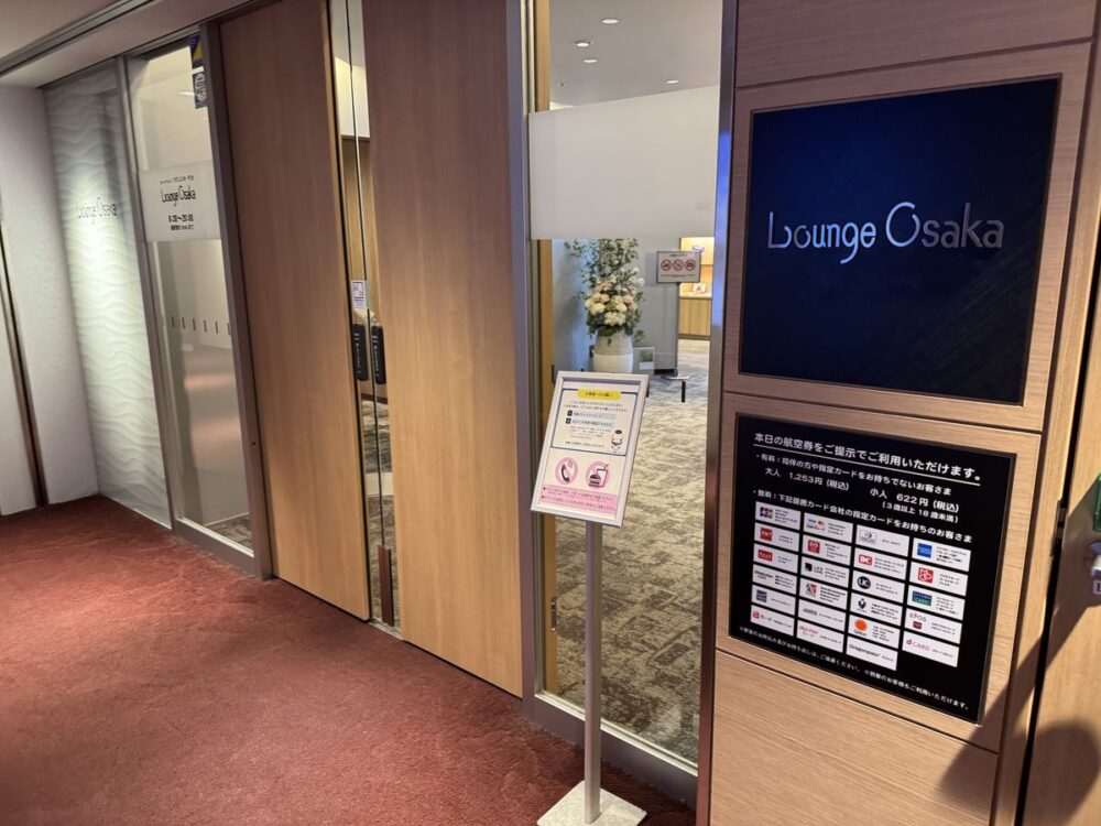 Lounge Osaka 入口の案内パネル、対象クレジットカードロゴ一覧と料金・営業時間表示、ガラスドア越しに内部の花瓶が見える