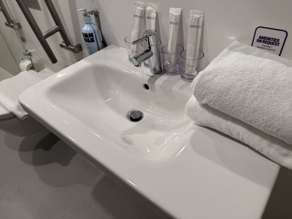 広い洗面台とたたまれたタオル。壁に「AMENITIES ON REQUEST」の案内